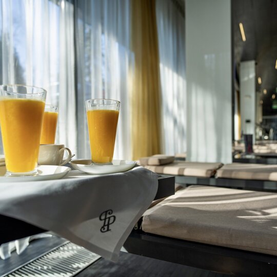 Trzy szklanki soku pomarańczowego i filiżanki na białej tacy w słonecznym hotelowym salonie z leżankami.