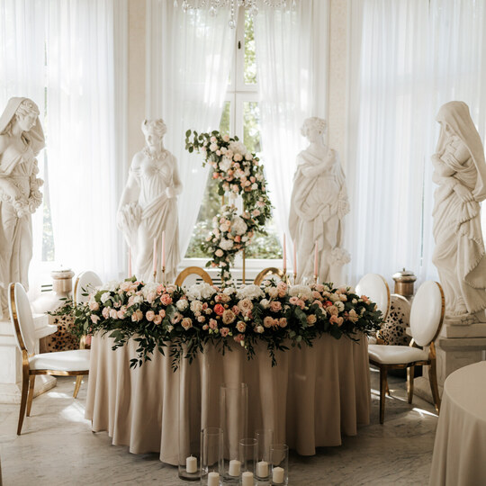 Elegant wedding head table with lush rose garland, tapered candles, gold-trim chairs and classical marble statues by sunlit windows.