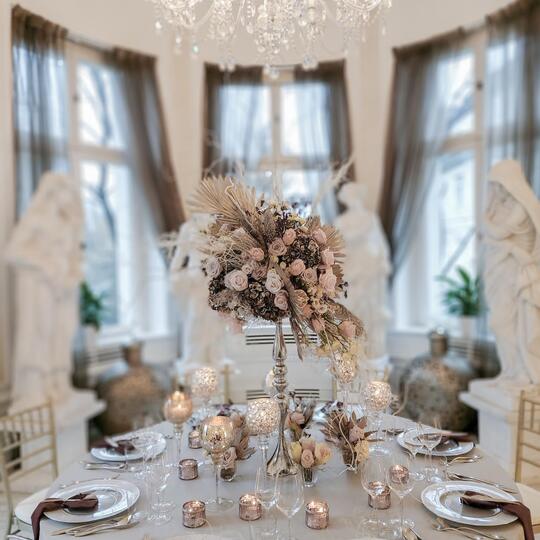 Elegant round dining table for formal dinner, crystal chandelier overhead, tall floral centerpiece, candles and place settings.