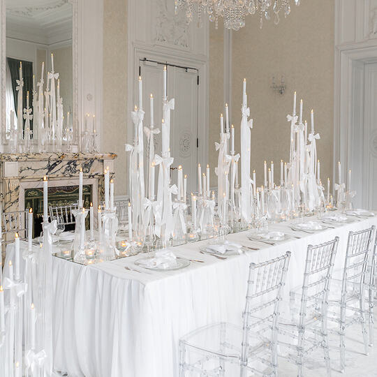 Elegant long banquet table with white linens, tall ribbon-tied candles, crystal chandelier and clear ghost chairs