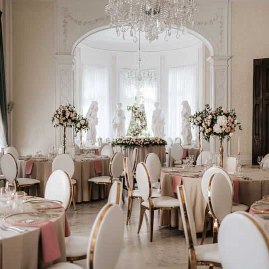 Elegant banquet hall with crystal chandeliers, ornate arch and statues, round tables with pink napkins, gold-trim chairs and tall floral centerpieces