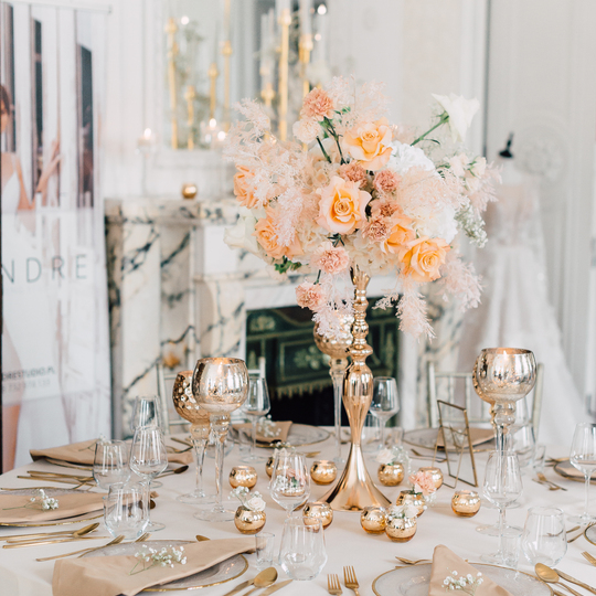 Elegant wedding table with tall gold vase, peach rose centerpiece, gold cutlery, glassware and marble fireplace