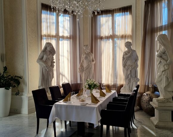 Elegantní soukromý jídelní salonek s krystalovým lustrem, mramorovou podlahou, klasickými sochami u erkeru a stolem pro osm