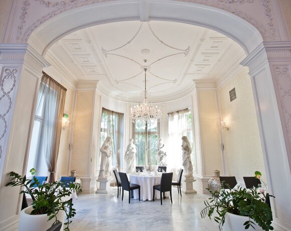 Elegant dining room with round table set under a crystal chandelier, classical statues, marble floor and tall arched windows.