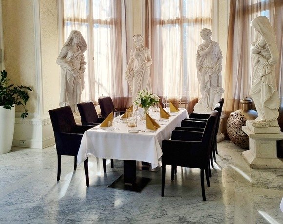 Elegancka prywatna sala jadalna z marmurową podłogą, kryształowym żyrandolem, rzeźbami i stołem dla ośmiu przy wysokich oknach