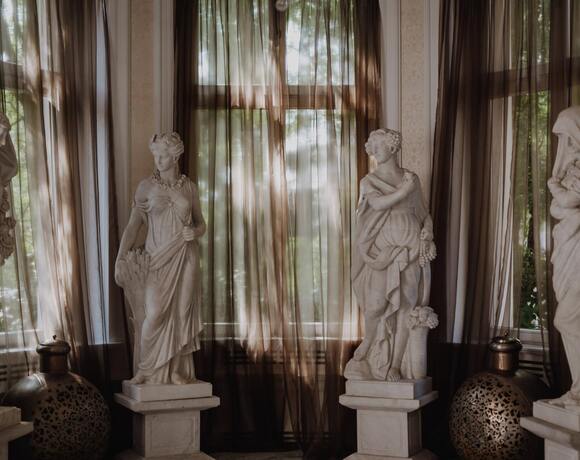 Hotel lounge with four classical marble statues on pedestals, tall windows with sheer brown curtains and decorative urns