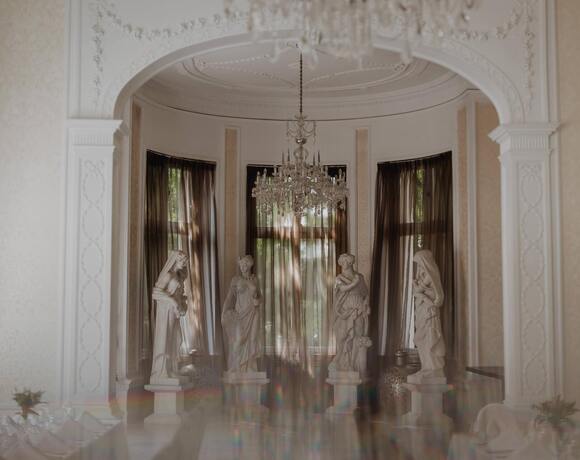 Elegant hotel dining room with ornate arch and chandelier, four marble statues by tall windows with sheer curtains and white‑linen tables.