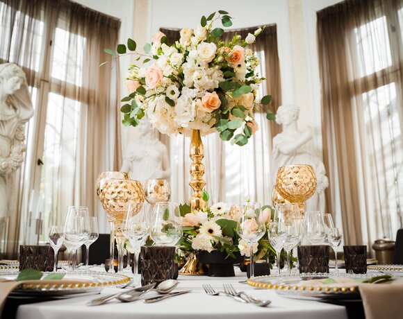 Elegancki stół bankietowy z wysoką kompozycją kwiatową, złotymi świecznikami, kryształowymi kieliszkami i marmurowymi rzeźbami przy drapowanych oknach