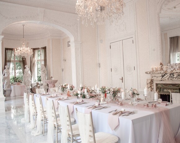 Elegant banquet room with long wedding table, pink runners, floral centerpieces, candles, crystal chandeliers and marble fireplace