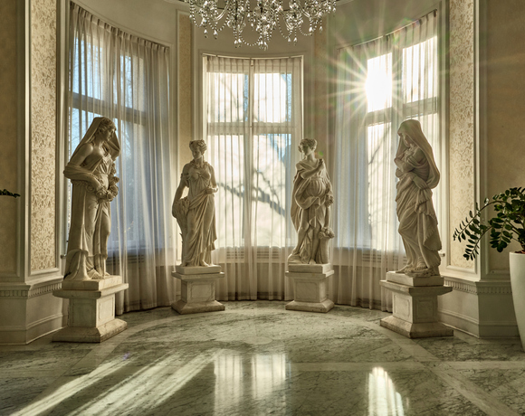 Foyer d'hôtel élégant avec sol en marbre, lustre en cristal, quatre statues classiques près de baies vitrées ensoleillées et plantes.