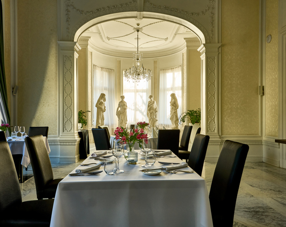 Elegant hotel dining room with white-tablecloth table, black leather chairs, crystal chandelier and classical statues in a sunlit bay