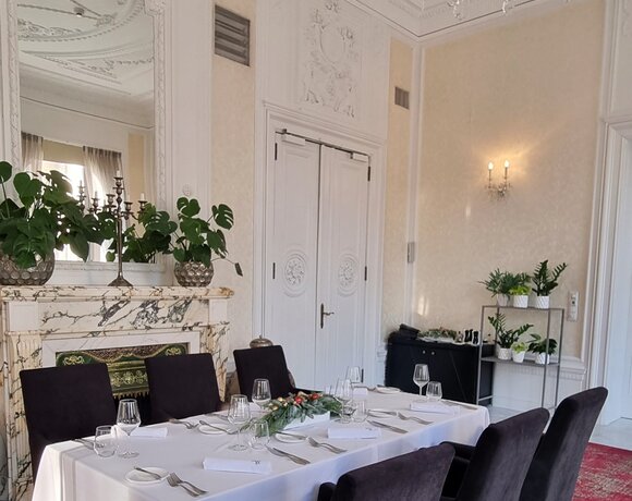 Elegant dining room with long white‑clothed table set for guests, velvet chairs, crystal chandelier, marble fireplace and ornate plaster ceiling