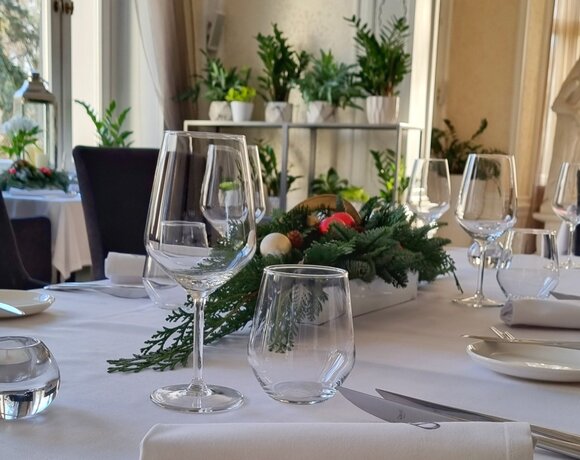 Elegant restaurant table with crystal wine glasses, rolled napkin, festive evergreen centerpiece and sunlit plants in background