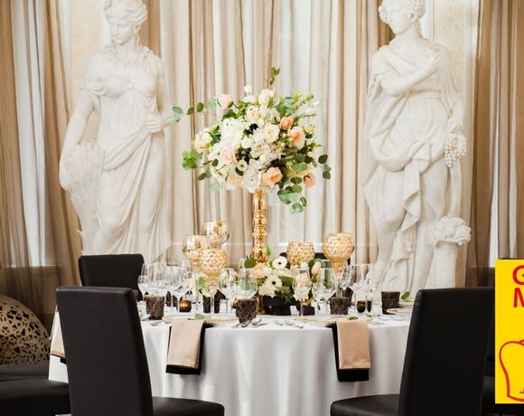 Elegant round table set for fine dining with gold floral centerpiece, crystal glassware, black chairs and classical statues backdrop