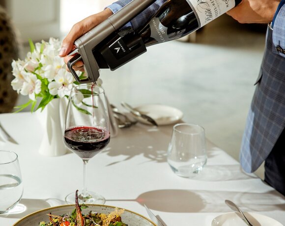 Sommelier pouring red wine into a glass at a white-tablecloth fine-dining table, plated steak with sauce and gourmet salad.