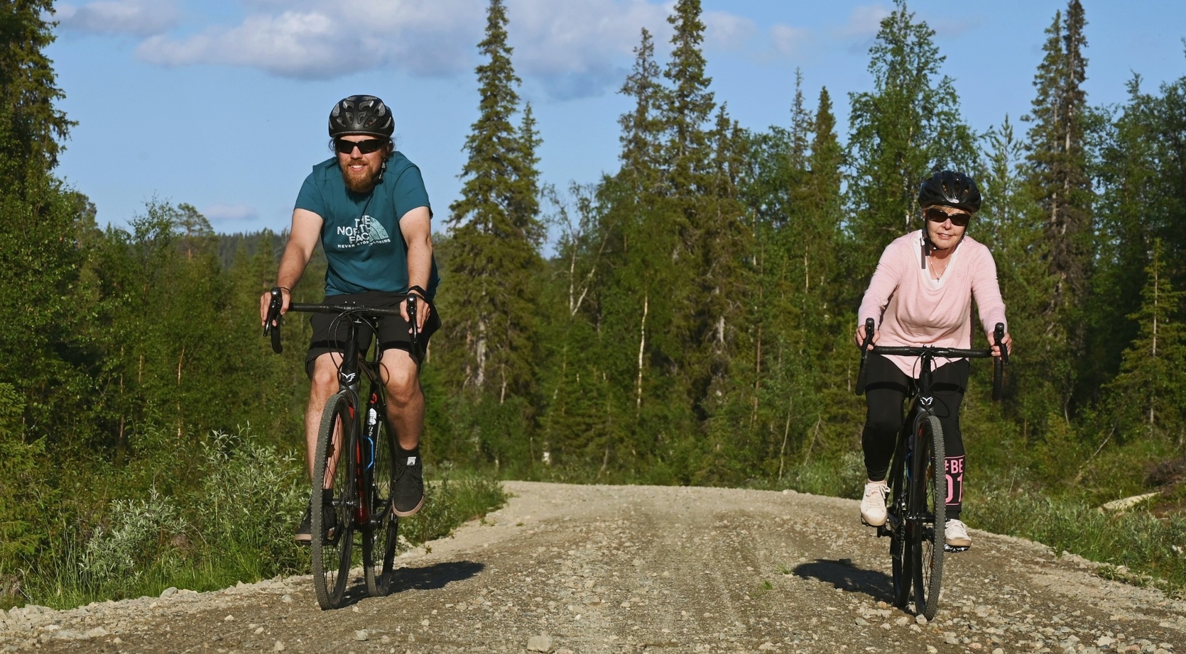 Two helmeted cyclists riding a sunlit gravel trail through a dense pine forest under clear blue skies