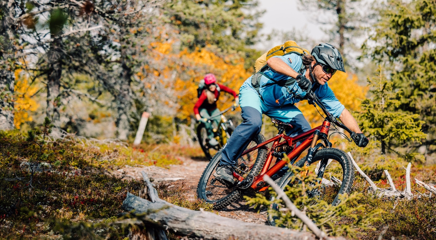 Two riders on a forest mountain-bike trail in autumn, guide-led adventure on full-suspension bikes with helmets and backpacks