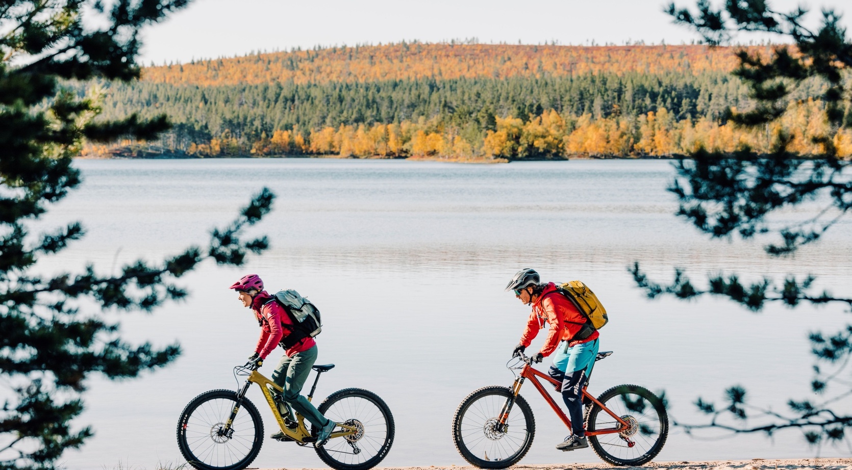 Two cyclists on a lakeside trail with backpacks, framed by pines and an autumn forest across calm water