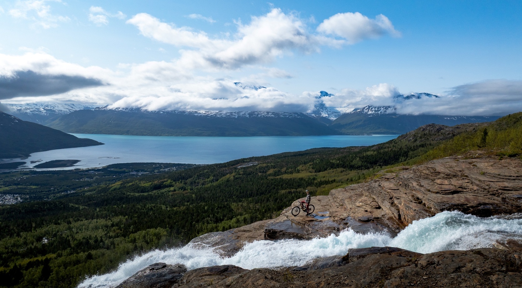 Cyclist on a rocky ledge beside a rushing waterfall, overlooking a turquoise fjord and snow‑capped mountains under blue sky