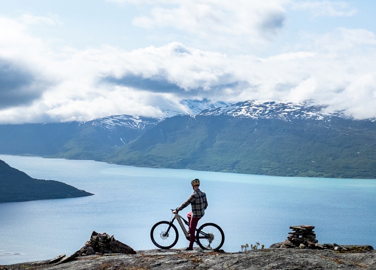 Cyclist with mountain bike on rocky overlook above turquoise fjord, snow-capped mountains and cloud-streaked sky