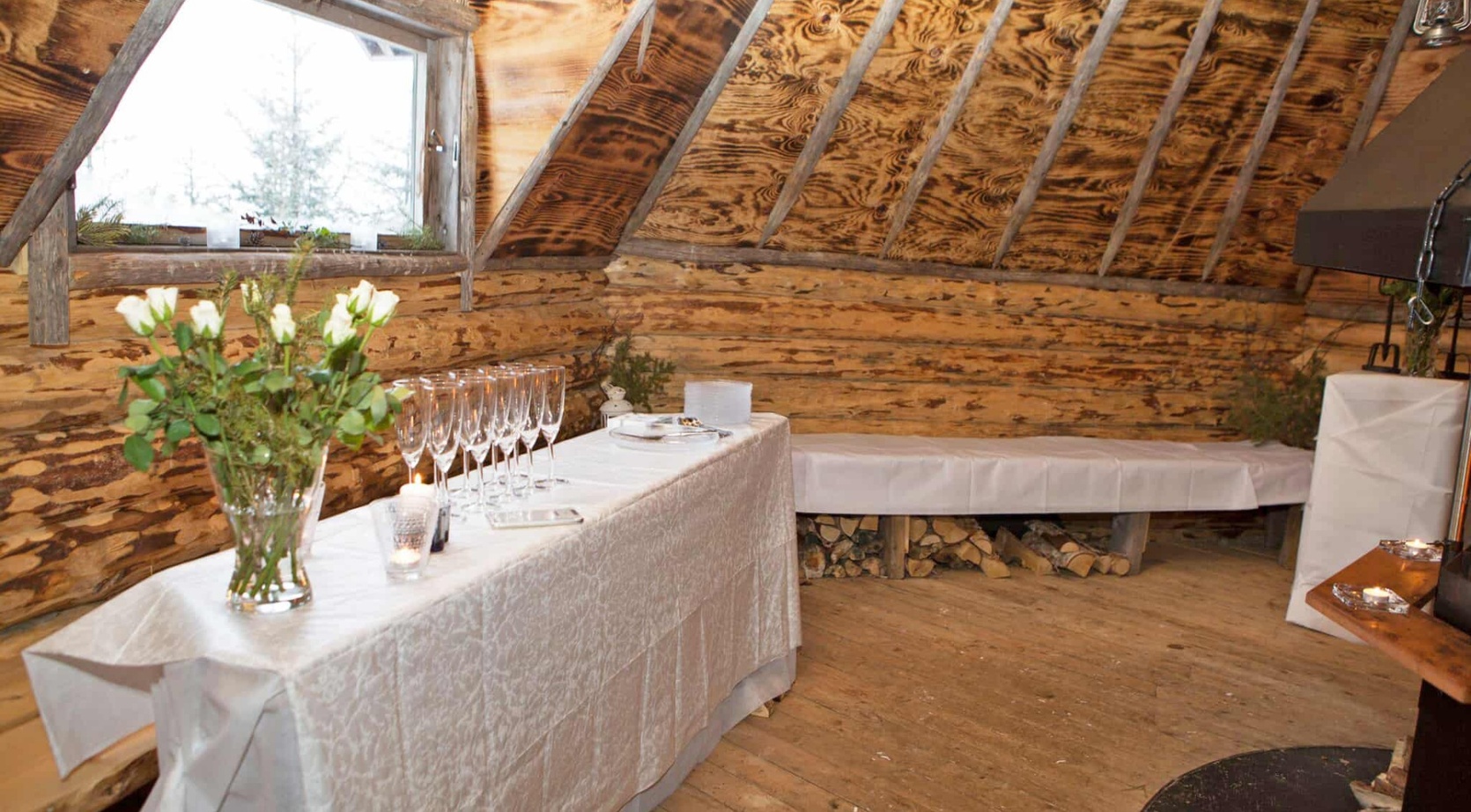 Rustic loft event room with sloped wood ceiling, white-clothed table set with champagne flutes, white roses, candles and bench over stacked firewood