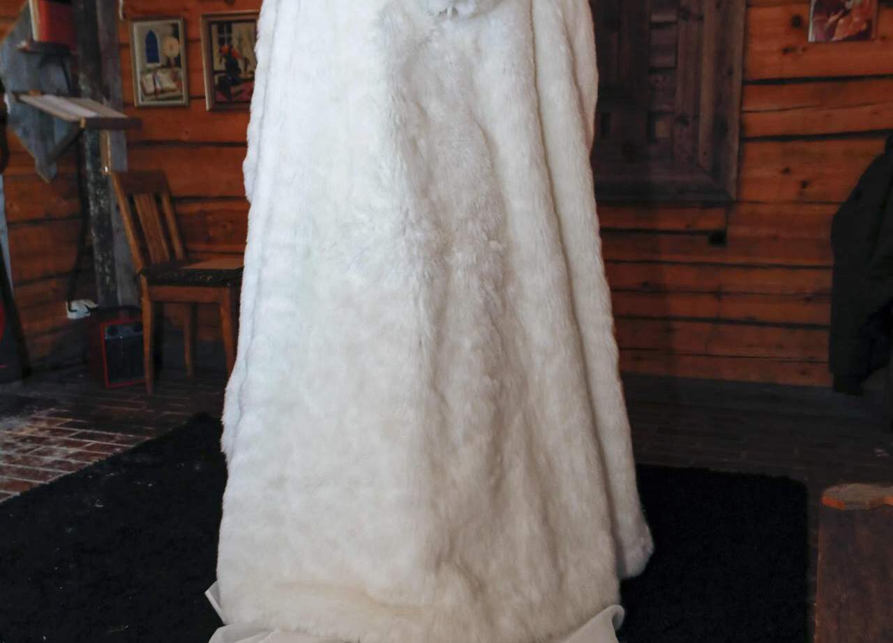 Bride standing with back to camera in rustic wooden chapel, wearing a white fur cloak over lace wedding train.
