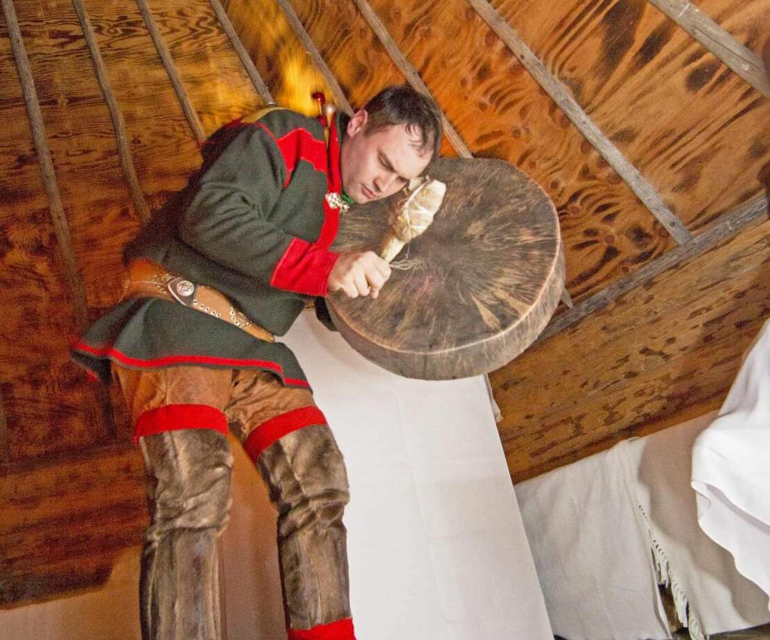 Performer in traditional Sámi dress playing a large frame drum inside a wooden hut, wearing fur boots.