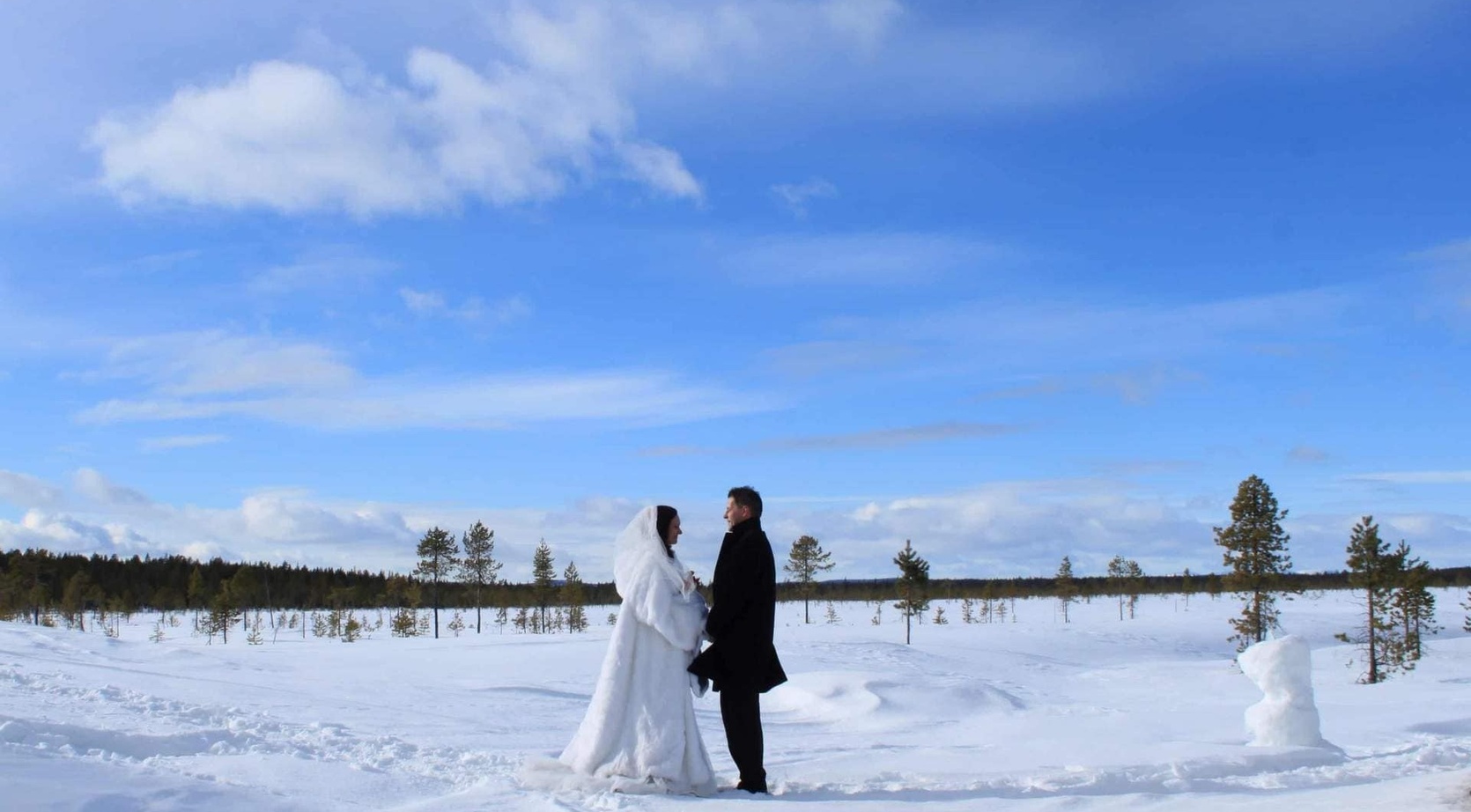 Couple in wedding attire holding hands on a snowy plain with scattered pines under a bright blue sky