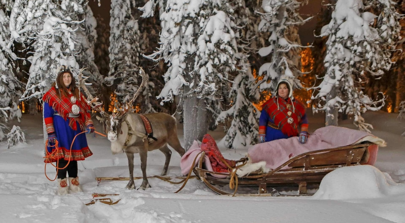 Two Sami hosts in colorful traditional dress with a reindeer and wooden sled draped in blankets in snow-covered forest