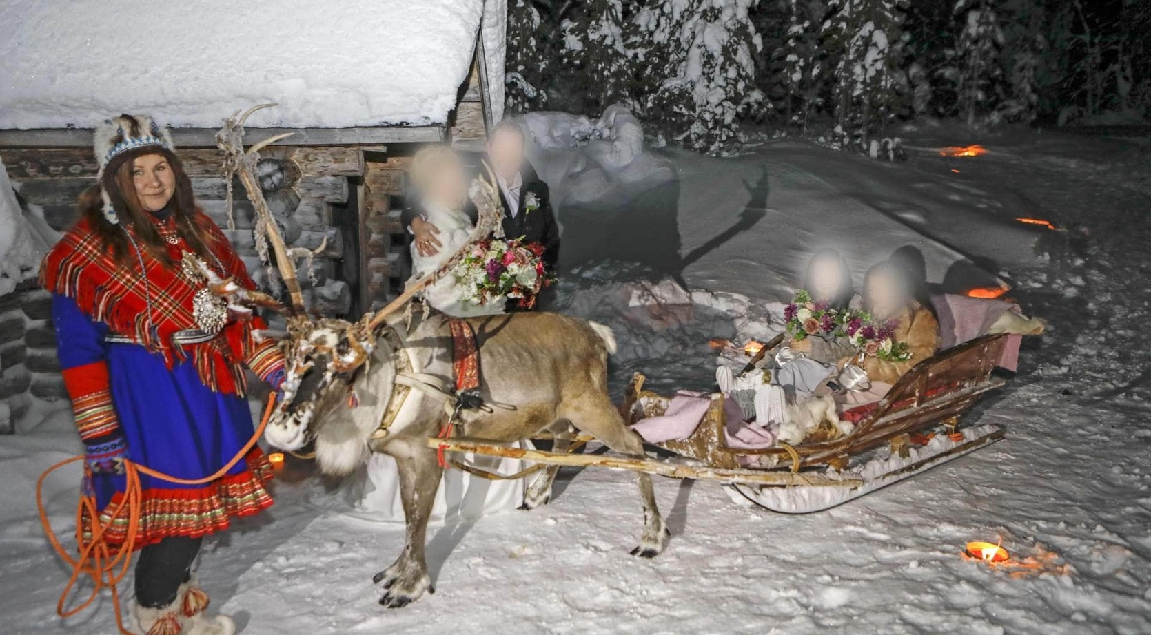 Reindeer sleigh ride at snowy lodge — Sami guide in traditional dress, blanketed guests holding bouquets on a candlelit trail.