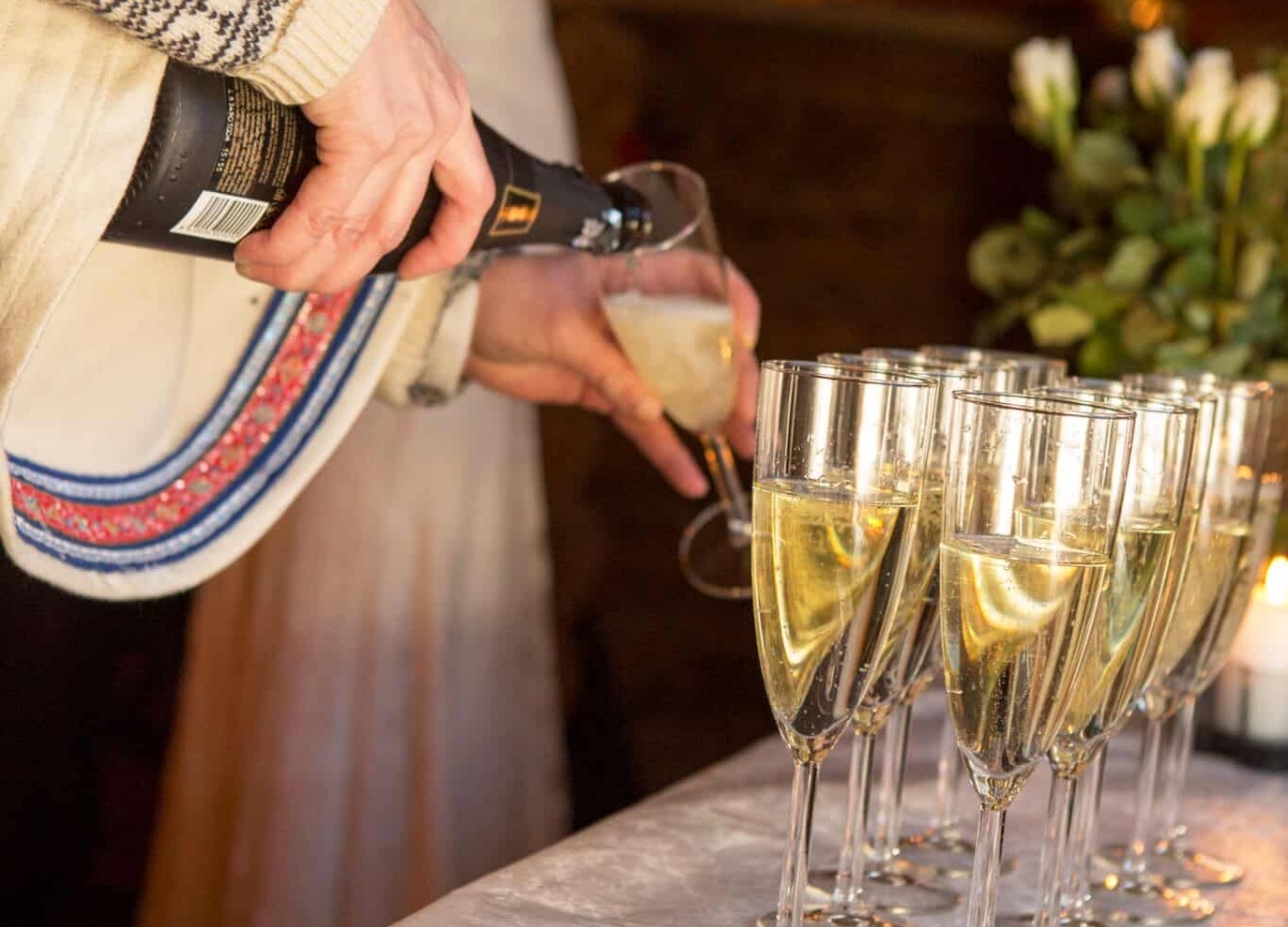 Person in embroidered jacket pouring champagne into flutes beside a chocolate cake and flowers, warm reception ambience