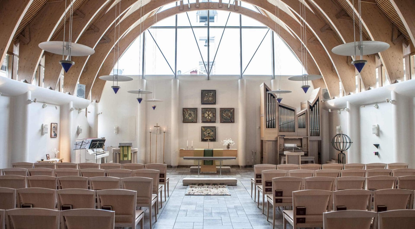 Bright chapel with soaring wooden arches, large arched window, pipe organ, simple altar and rows of pale wooden chairs.