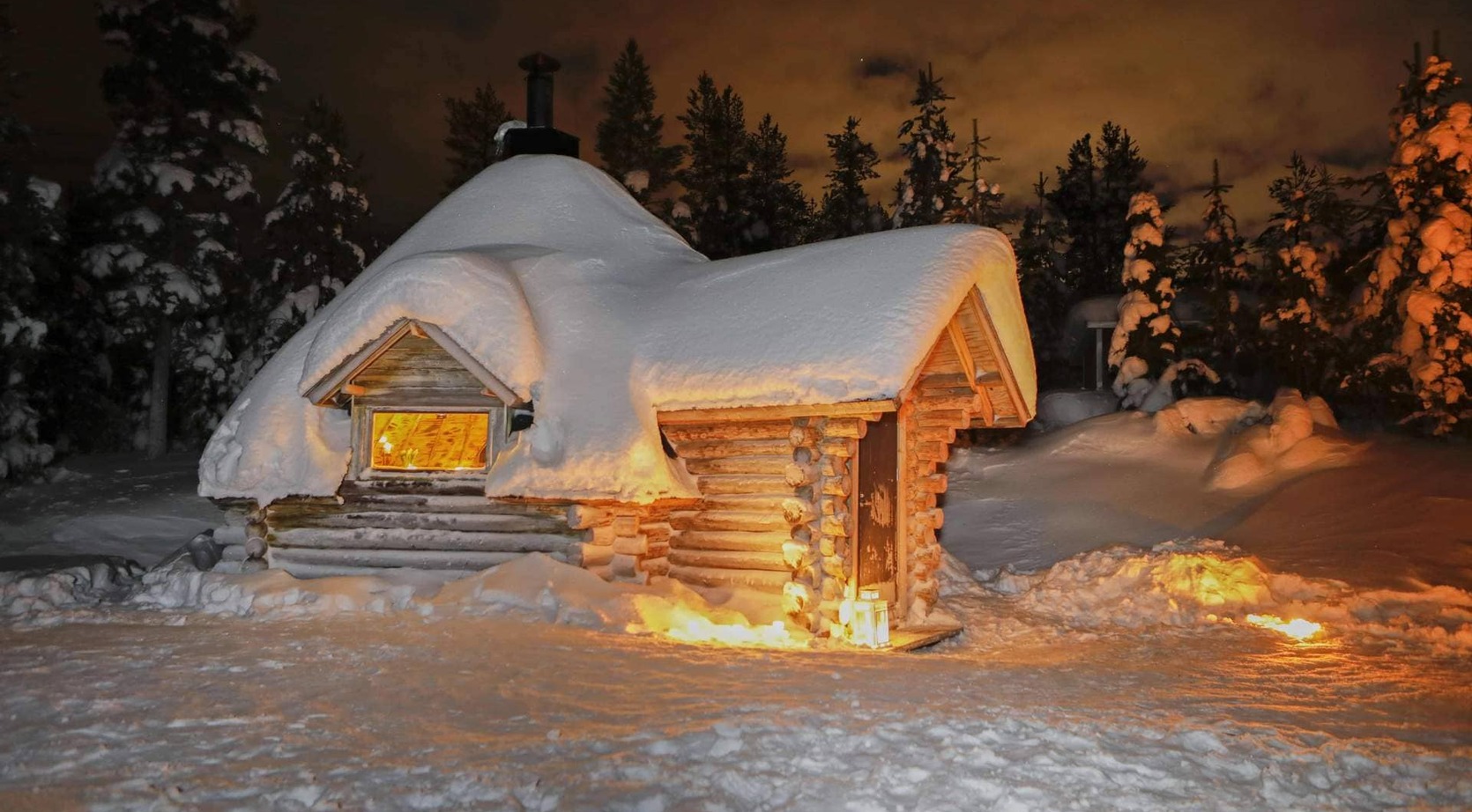 Snow-covered log cabin with thick roof drift, warm glowing window and lanterns, secluded forest setting at night