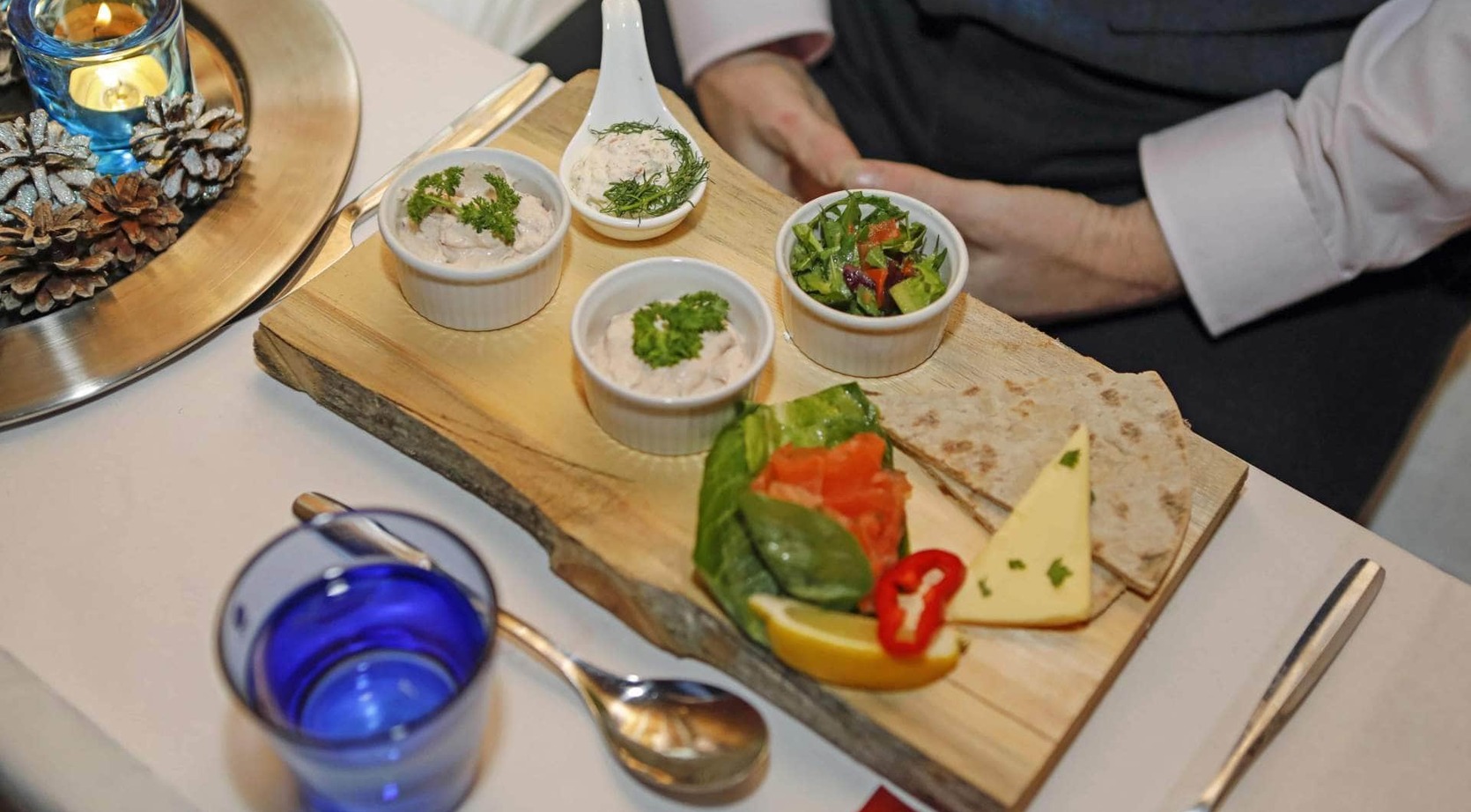 Wooden appetizer board with dips in ramekins, small salad, smoked salmon, cheese, flatbread and lemon wedge and a lit candle.