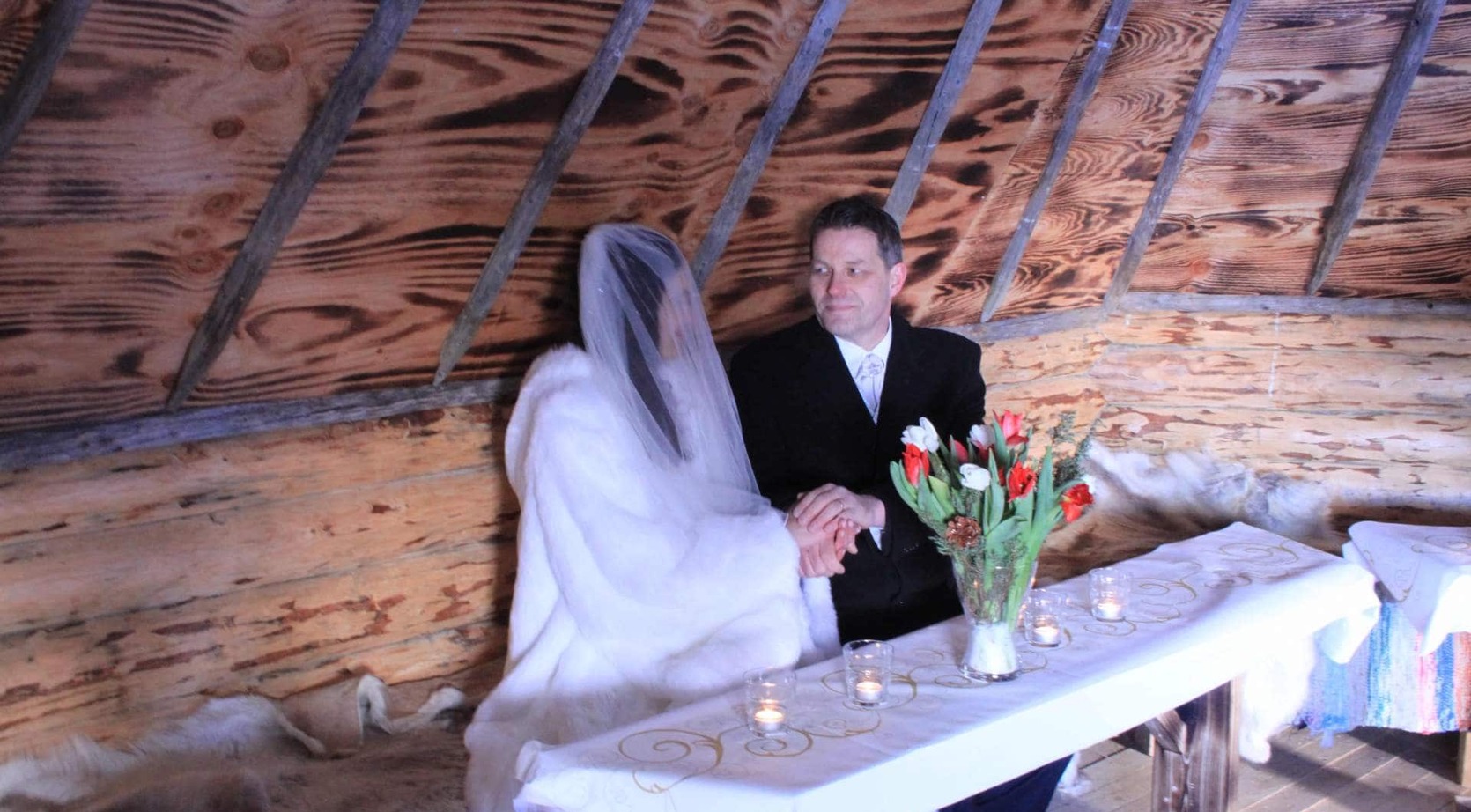 Bride and groom holding hands at a candlelit table in a cozy rustic log-cabin setting with fur throws and tulip bouquet