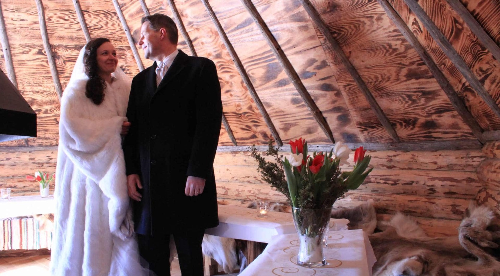Smiling bride and groom in winter coats at an intimate rustic cabin wedding, fur-lined benches, tulip centerpiece and wooden-beamed ceiling.