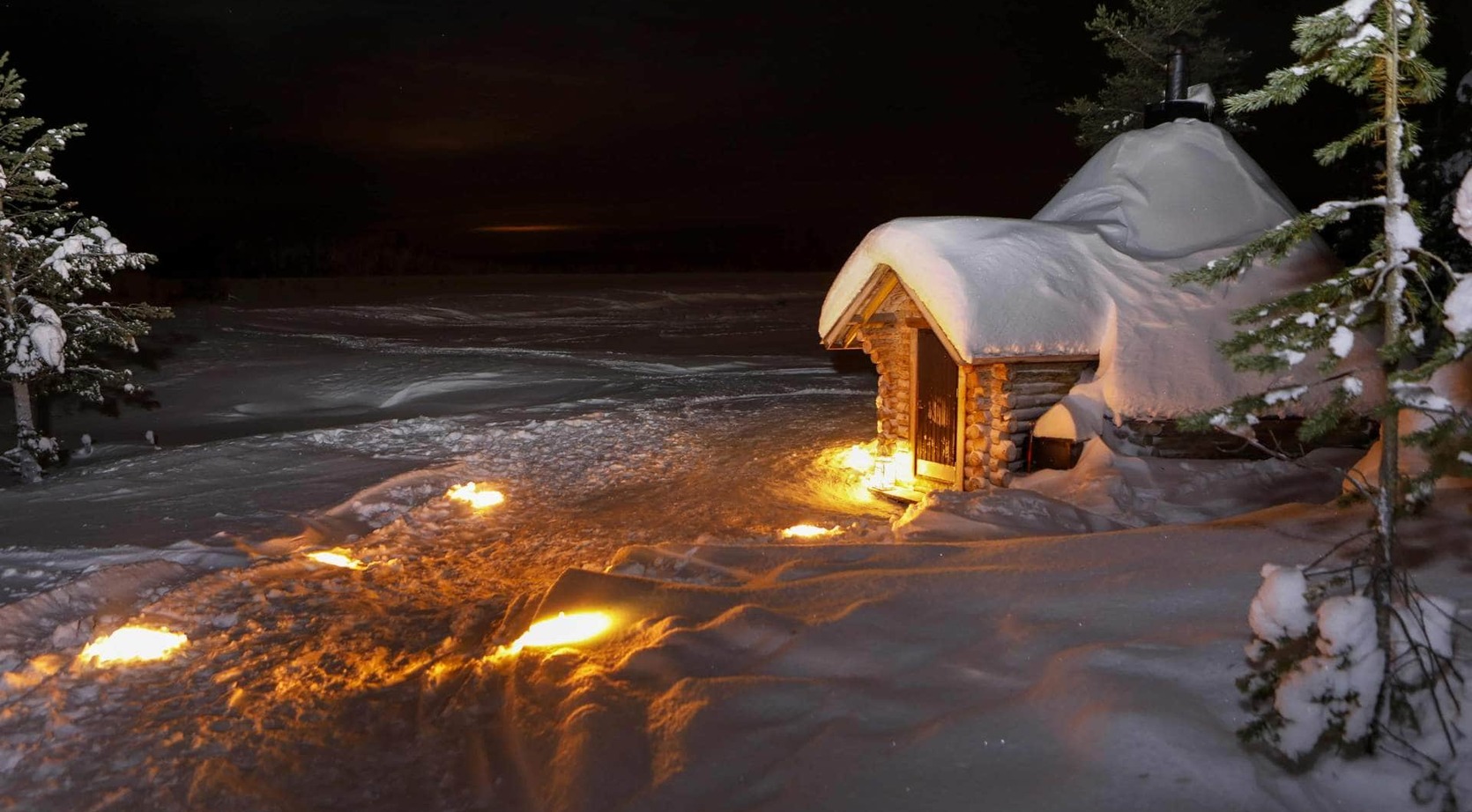 Snow-covered wood sauna by a frozen lake at night, torch-lit path and snow-laden pines creating a cozy winter ambiance