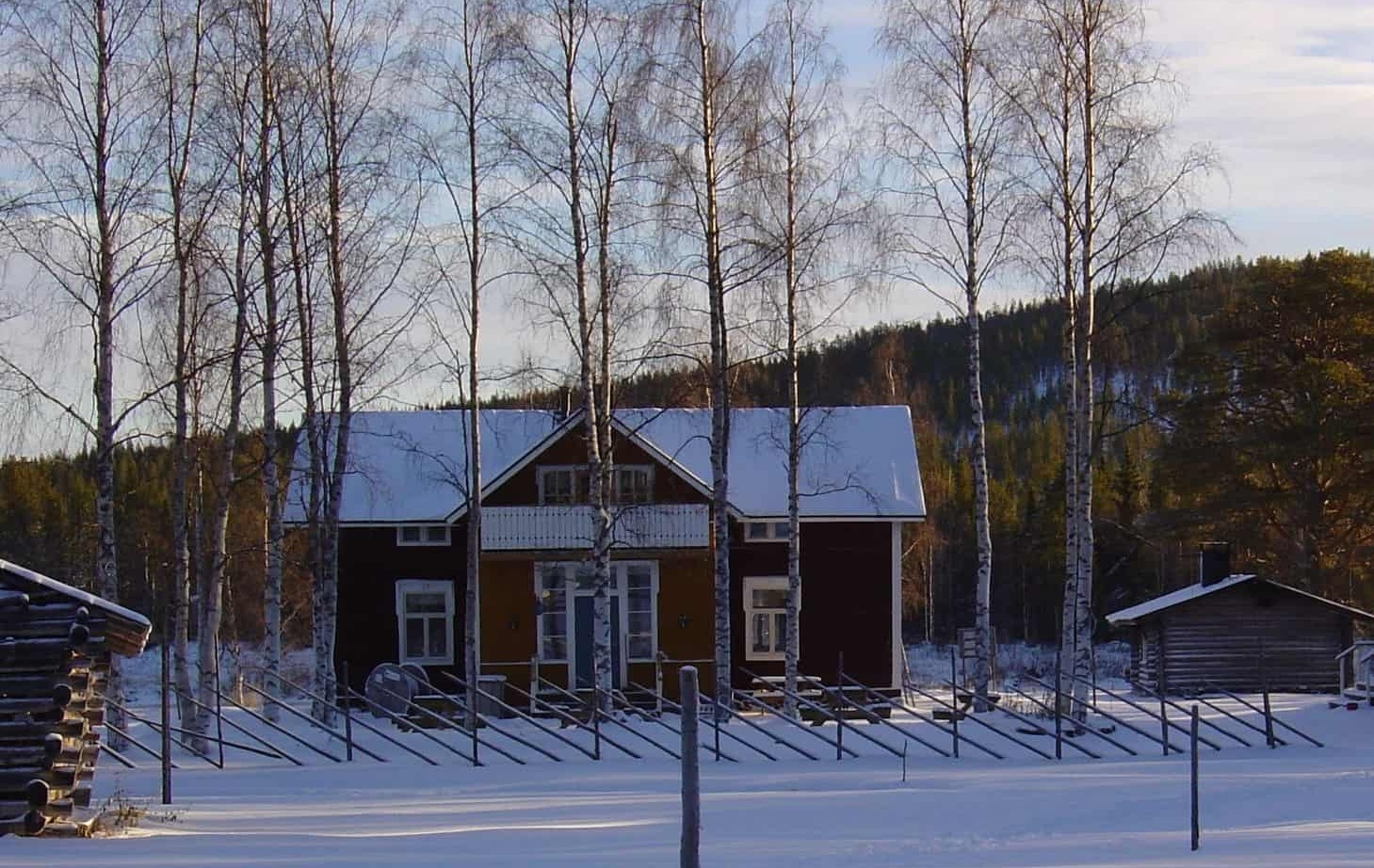 Red wooden country lodge with snow-covered roof, birch trees and rustic drying racks, log outbuilding and forested hill backdrop.