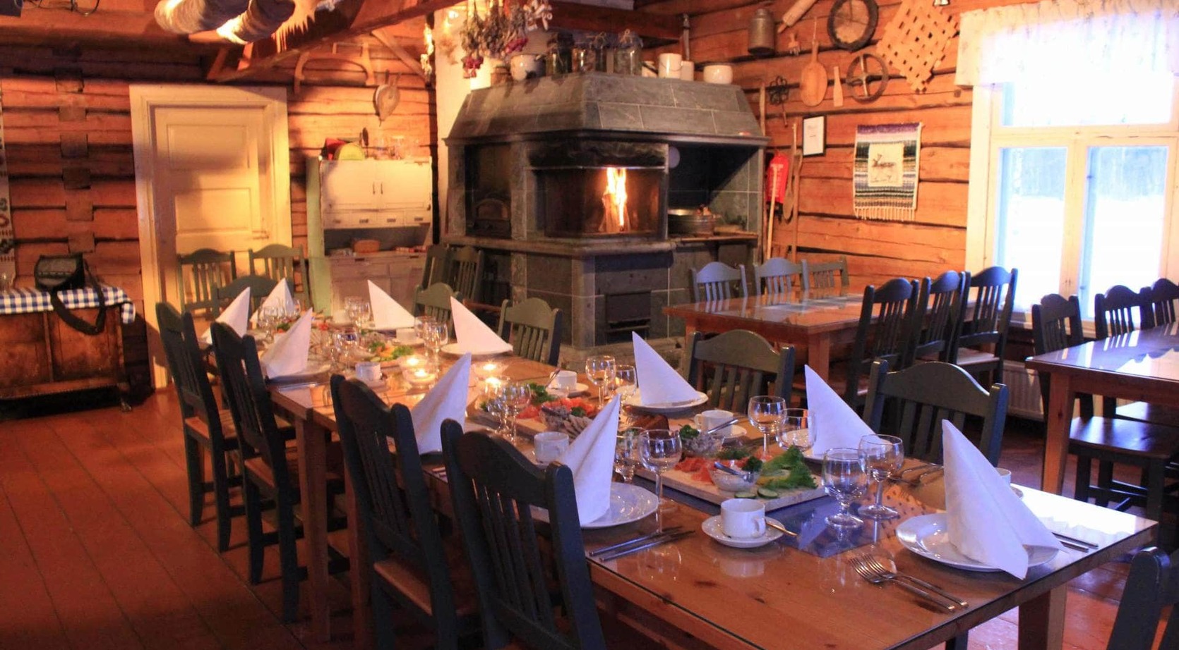 Rustic log cabin dining room with long wooden tables set for a communal meal, napkins, cozy stone fireplace and warm light