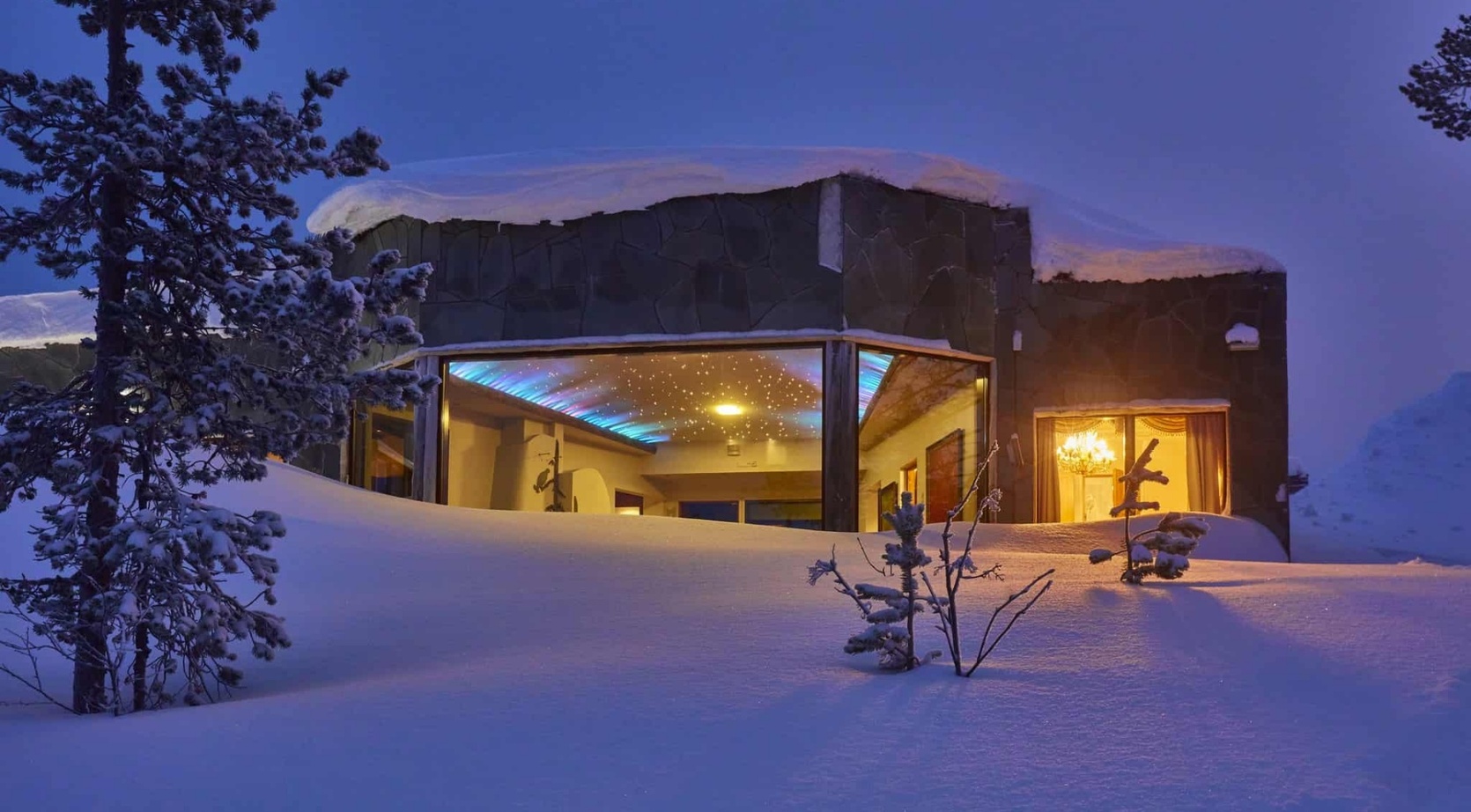 Snow-covered modern hotel with panoramic windows revealing a warm lounge with starry ceiling lights and chandelier