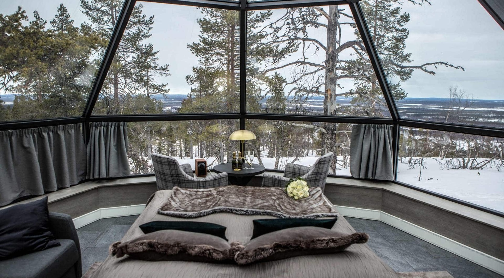 Panoramic glass suite with king bed, fur throw, two plaid chairs and table set with champagne, snowy pine forest view