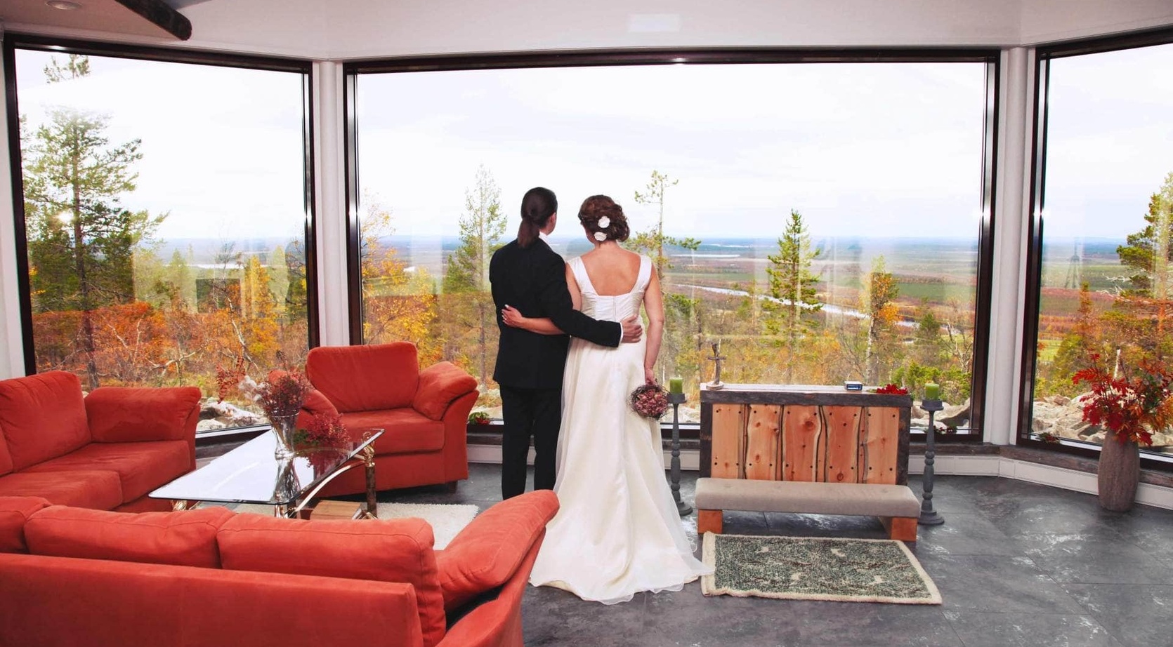 Bride and groom embrace at panoramic floor-to-ceiling windows in a cozy lounge with red sofas, overlooking autumn forest.