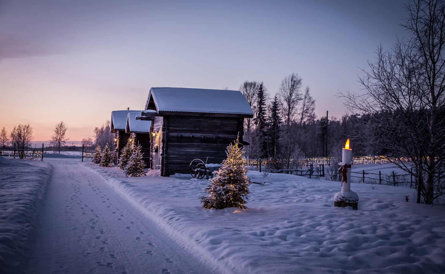 Snow-covered wooden cabins at twilight, decorated with lit Christmas trees and a glowing candle by a snowy path