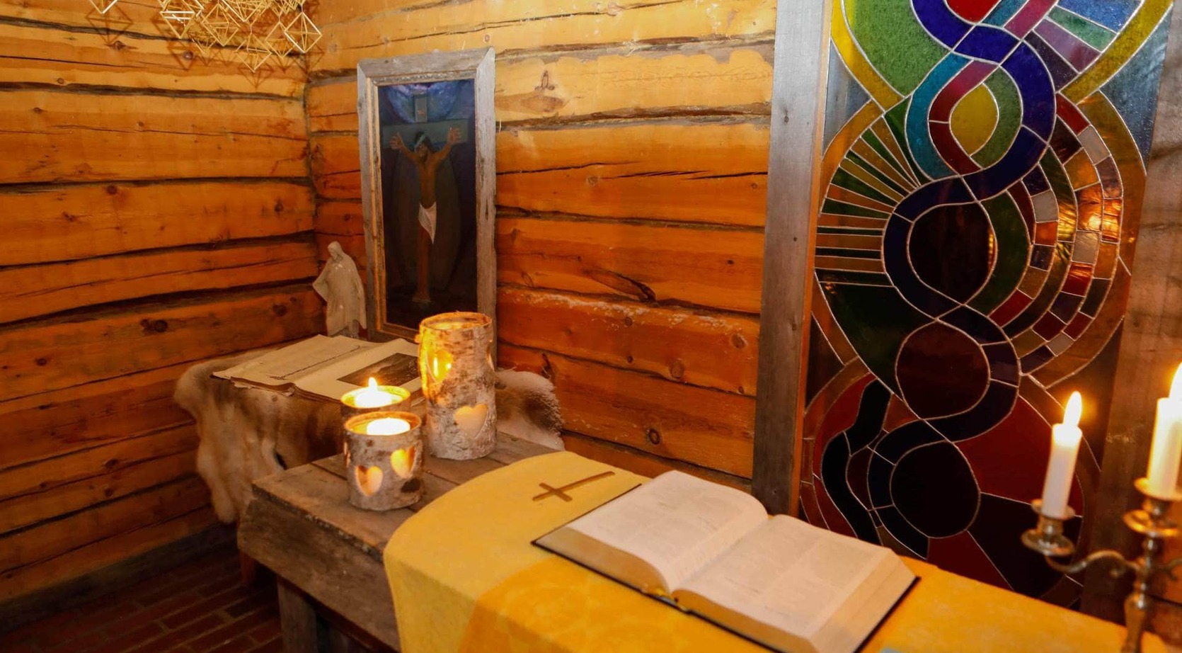 Cozy rustic chapel corner with log walls, small altar, open Bible, lit birch-candle holders and colorful stained-glass window