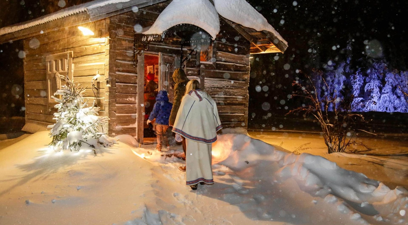 Snow-covered log cabin with a warmly lit entrance as guests in winter coats and a decorative cloak enter during heavy nighttime snowfall