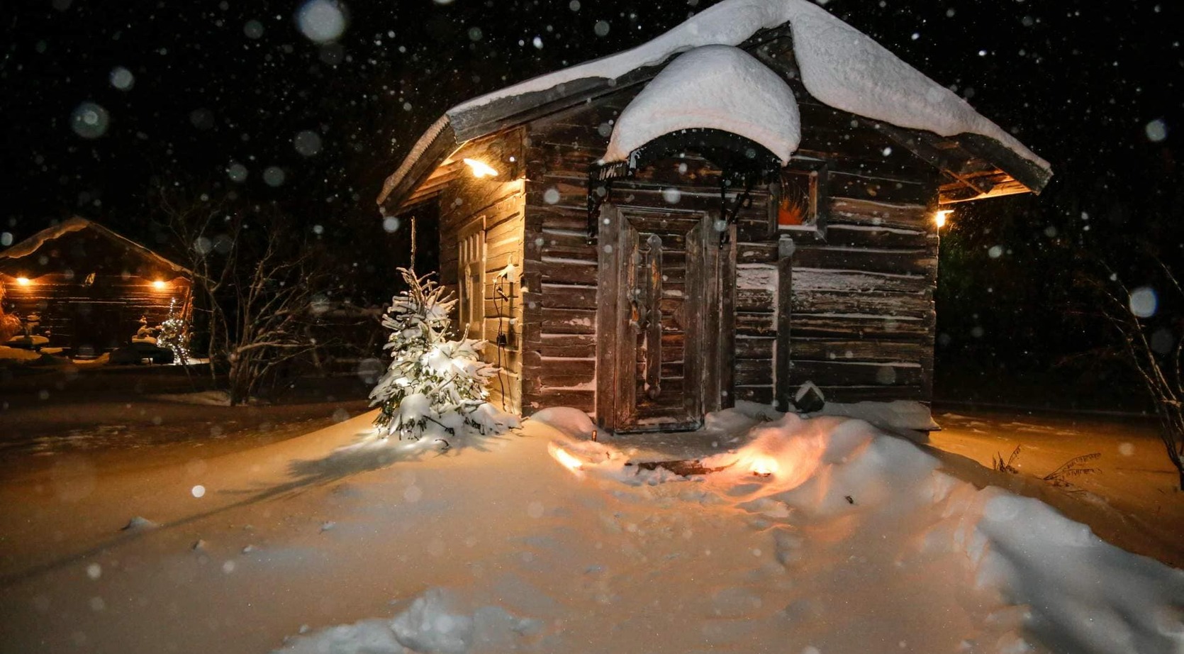 Cozy snow-covered log cabin at night, warm exterior lights, small decorated spruce and heavy snow piled on curved roof.