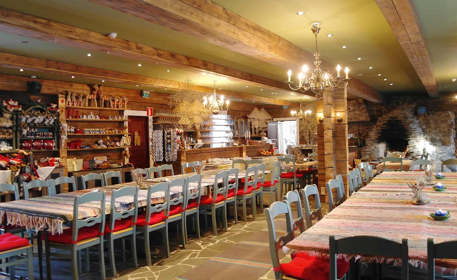 Rustic hotel dining room with long communal tables, wooden beams, chandeliers, stone fireplace, woven runners and red seat cushions.