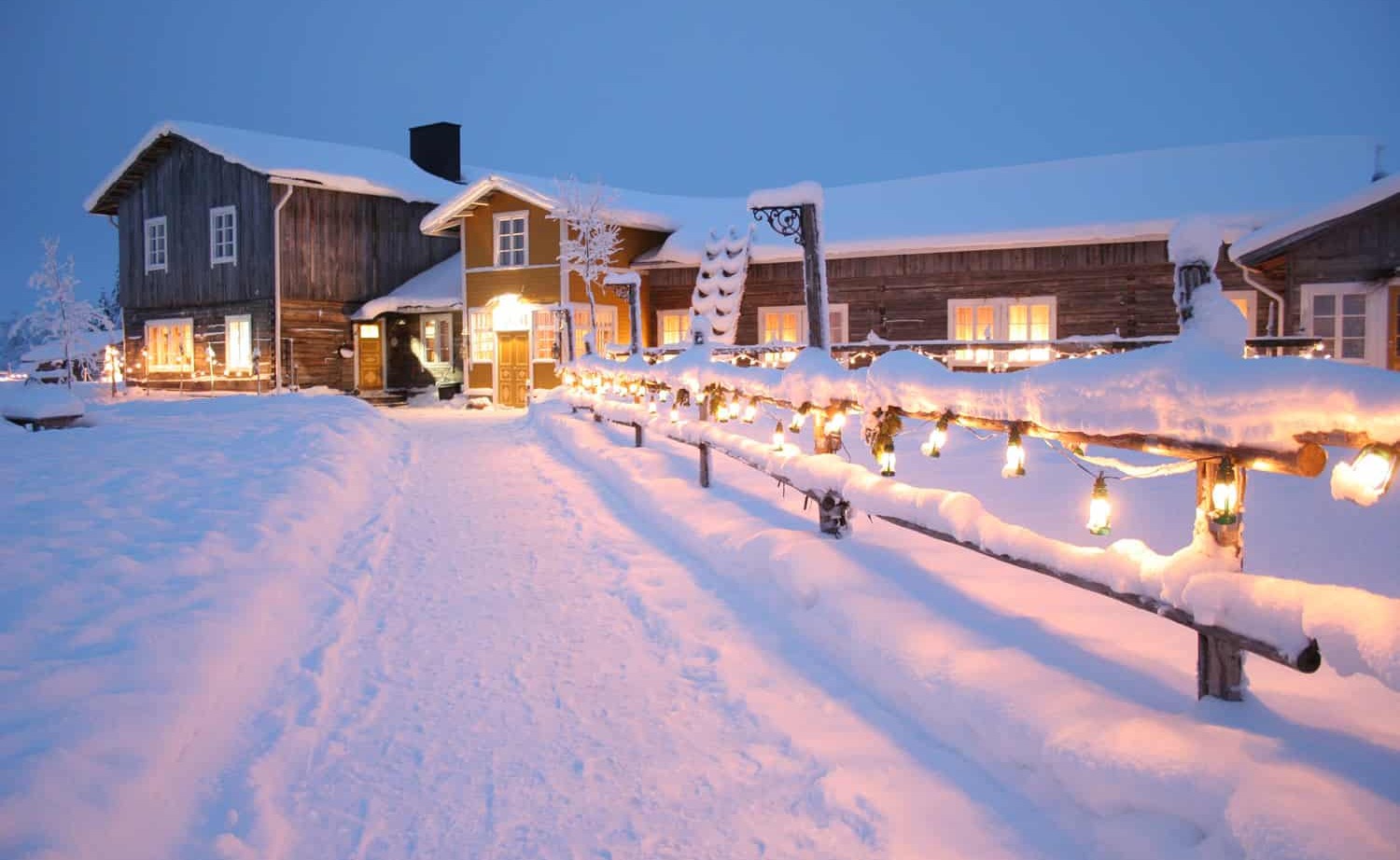Rustic snow-covered lodge with glowing windows and lanterns lining a snowy fence path to the warmly lit entrance