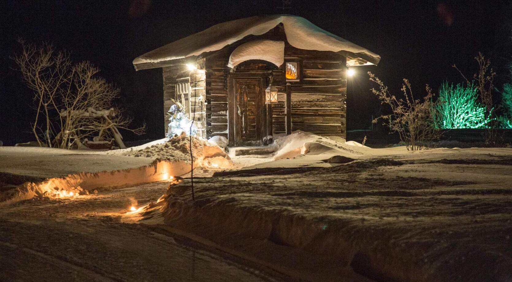 Snow-covered log cabin at night, warmed by lanterns and a candle-lit path, cozy wooden entrance and frosted shrubs