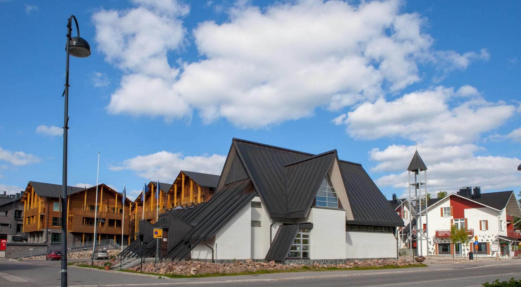 Chalet-style wooden hotel and modern steep-roofed lodge beside a bell tower under a bright blue sky with soft white clouds